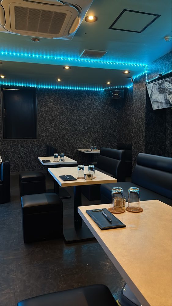 青い照明が天井に光る、スナックCloverの店内。白いテーブルと黒いソファが並ぶ